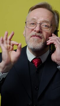 Happy senior businessman talking the phone. Portrait of a mature man in black costume speaking on the mobile phone and gesticulating a lot isolated on yellow. Vertical video