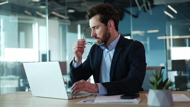 Lamentable caucasian male in formal attire feeling headache in modern office while working on wireless laptop. Well-lit spacious and contemporary environment. Concept of pain and technology.