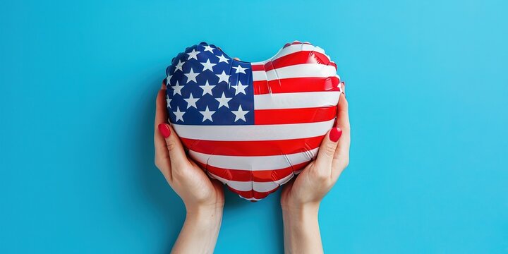 A pair of hands holding an American flag heart-shaped balloon, rising against a clear blue backdrop - Powered by Adobe