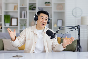 Enthusiastic Asian podcaster talking with hands, having an engaging conversation indoors, looking at camera with headphones and microphone.