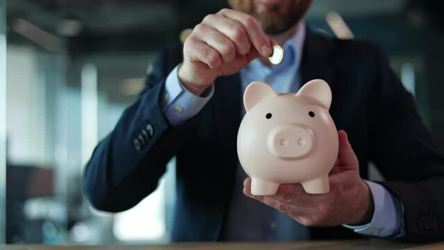 Close up of unknown bearded male in business suit inserting coin in piggybank while sitting by table. Provident economy manager presenting idea of finances collecting and saving money for future.