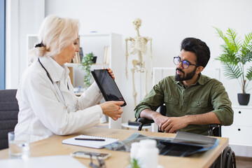 Fototapeta na wymiar Attractive bearded young man looking on tablet with x-ray image carried by radiologist in consulting room. Confident senior specialist discussing results of diagnostic tests with wheelchair user.