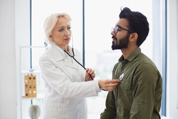 General practitioner examining heartbeat while providing regular checkup in clinic. Side view of focused elderly lady in white coat using stethoscope while young man sitting on exam couch. - Powered by Adobe