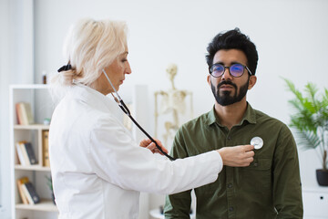 Serious bearded man in everyday wear visiting family doctor for full examination in general practice. Elderly woman in white lab coat checking lungs and heart via stethoscope.