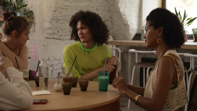 Homosexual people talk and drink cocktails at table in bar. Young lesbian women, gay man talking and drinking alcohol drinks, celebrating and sitting at desk in interior. Beautiful multiethnic friends