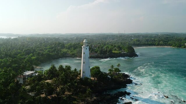 Aerial view of Dondra lighthouse on Sri Lanka coastline. Historic tower stands against blue ocean backdrop, waves crash on rocks. Coastal landscape from above with palm fringed shores.