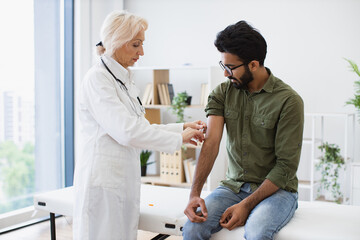General practitioner giving injection to boost immunity for young bearded patient. Confident mature woman in lab coat holding syringe with seasonal flu vaccine while young man sitting in exam room. - obrazy, fototapety, plakaty