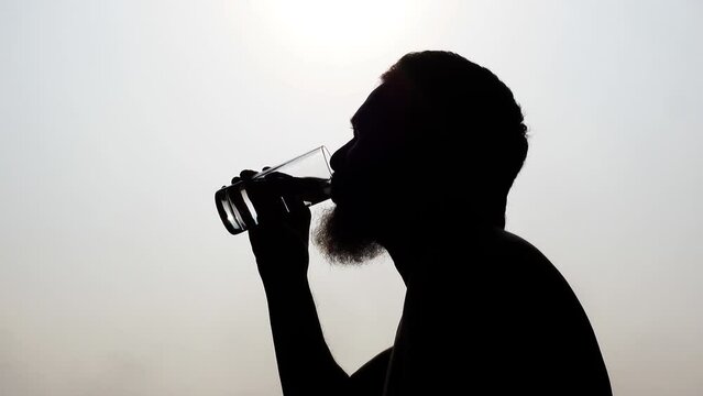 A thirsty old man drinks water from a transparent drinking glass in the hot sun during the summer midday. 