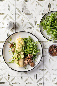 French rolled omelette stuffed with cream cheese with avocado, aragula and mushrooms for breakfast on white plate printed tile background, top view