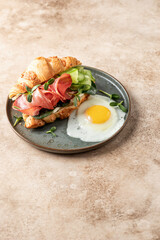 Fresh croissant sandwich with prosciutto or ham, cucumber, soft cheese and micro green and fried egg for breakfast isolated on beige background