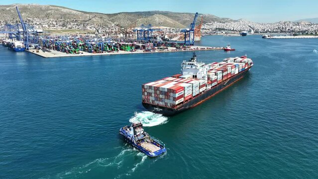 Aerial drone video of container cargo vessel approaching logistics unloading import and export container terminal of Perama - Piraeus assisted by tug boat