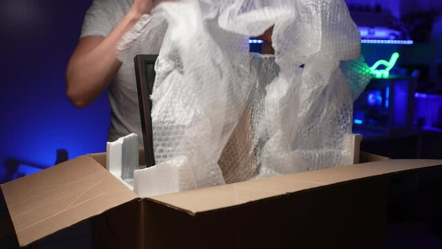 Cropped front view of unrecognizable man unpacking cardboard box with new gaming monitor wrapped in plastic wrap and foam, standing by desk in dark room with blue neon light on background.