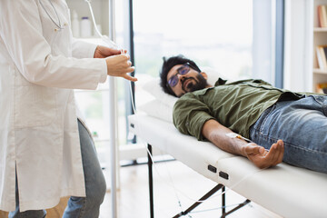 Cropped view of efficient general practitioner checking solution for chemotherapy of male lying on exam couch. Young bearded man in casual wear taking dropper while mature female making system review.