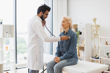 Side view of focused adult man in white coat using stethoscope while elderly woman sitting on exam...