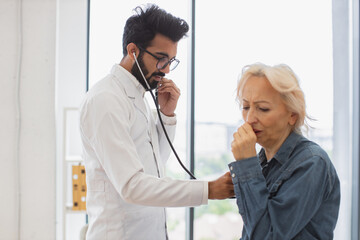 Serious old woman in everyday wear, coughing while visiting family doctor for full examination in general practice. Bearded man in white lab coat checking lungs and heart via stethoscope.