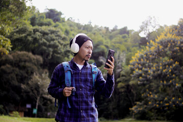 Young Asian man in a casual outfit, sporting a beanie, plaid shirt, and headphones, checks his phone with a puzzled expression on his morning nature walk. A backpack rests comfortably on his shoulder.