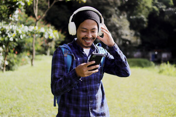 Young Asian man in a casual outfit, sporting a beanie, plaid shirt, backpack, and headphones, browses his phone for music, seemingly choosing the perfect playlist or song for his morning nature walk.