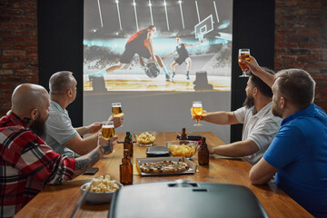 Men, friends gathering to watch online basketball match with enthusiasm translation on TV, drinking...