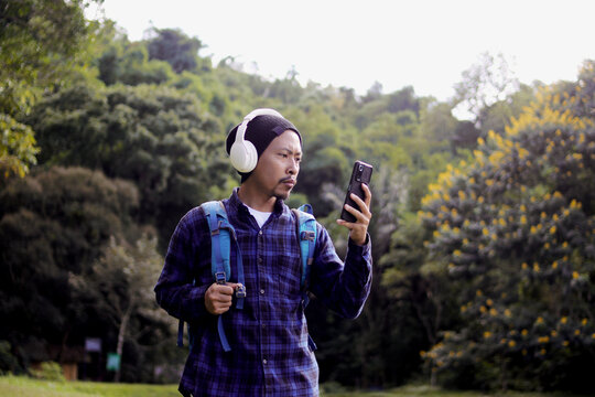 Young Asian man in a casual outfit, sporting a beanie, plaid shirt, and headphones, checks his phone with a puzzled expression on his morning nature walk. A backpack rests comfortably on his shoulder.