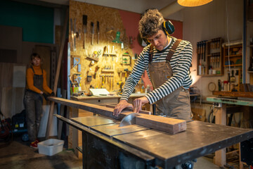Concentrated man carpenter wear noise cancellation safety headphones saws wood with circular saw in joinery workshop, female colleague helps hold the board. Woodwork, DIY, family small business