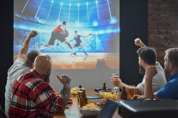 Obraz premium Men, friends sitting at table with beer and snacks, watching online basketball game translation on TV and cheering up favorite team. Concept of sport, championship, game, sport fans, leisure