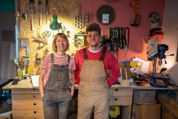 Relaxed crafts man and woman stands in carpentry room near table with tools, smiling looking at...
