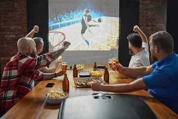 Men, friends sitting at table with beer and snacks, watching online basketball game translation on...