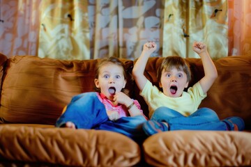 A young boy and girl of European appearance sit on a couch with their arms confidently raised in the air, expressing joy and excitement
