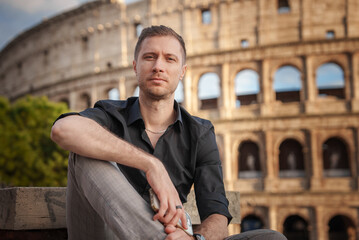 Man in black shirt and gray trousers sits at Colosseum in Rome with thoughtful expression. Iconic...