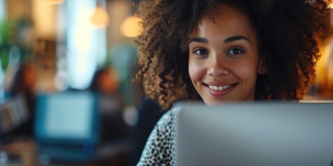 A woman with curly hair is smiling at the camera while holding a laptop
