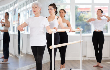 Fototapeta na wymiar Concentrated graceful elderly woman practicing basic ballet moves at barre during multi-age group class under guidance of experienced female instructor