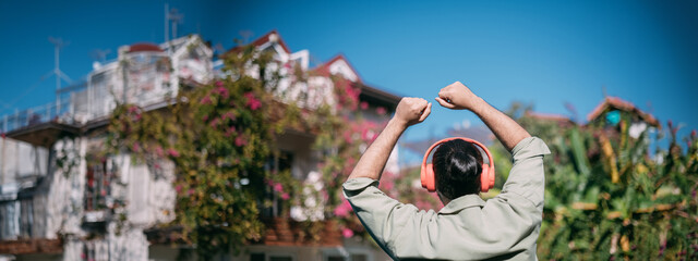 Portrait of a cheerful dancing young man in bright large headphones on a sunny day. A handsome guy in a green shirt happily dances, listens to music in a sunny garden