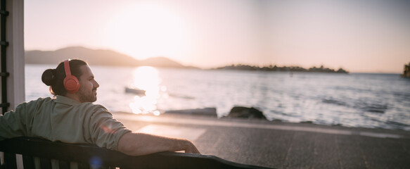 Portrait of a young man in bright large headphones listening to music, audiobook by the sea at sunset. - obrazy, fototapety, plakaty