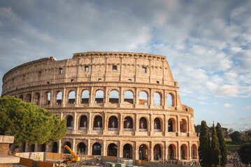 Fototapeta na wymiar Colosseum in Rome, Italy. Iconic ancient amphitheater under soft, cloudy daylight. Arch shaped ruins with construction work, tourists, and trees nearby.