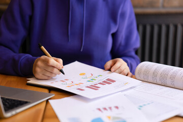 Female hands making notes on sheets with graphs and data with pen. Girl with blue sweater student working on assignment, business, freelancing, student in class, laptop on table, scientific graphs and