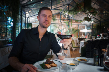 Elegant man in black shirt enjoys red wine at luxurious restaurant table with well presented meal and upscale dining ambiance. Modern, elegant interior.