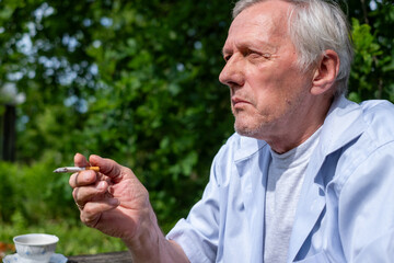 Deep in thought, a senior individual, elderly man, pauses for a smoke break outside, amidst the...