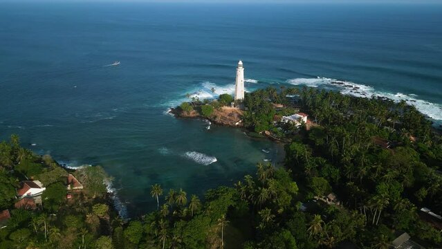 Aerial drone of Dondra lighthouse surrounded by palm trees in Sri Lanka. Waves crash against rocks, boats navigate nearby waters, serene beauty attracts travelers seeking picturesque coastlines.