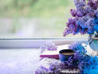 Obraz na płótnie Canvas A blue ceramic coffee cup and a bouquet of blooming lilacs on the background of a spring window. Copy space