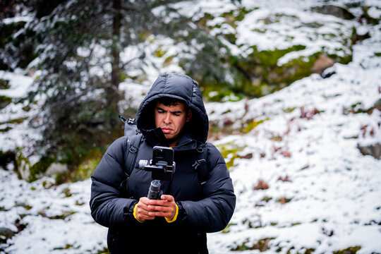 Young man uses mobile phone and gimbal to film snowy forest during snowfall.