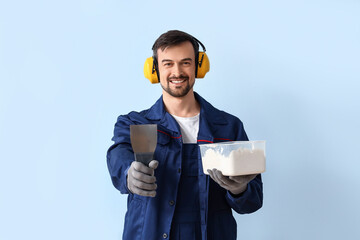 Male decorator in hearing protectors with putty knife and plaster on blue background