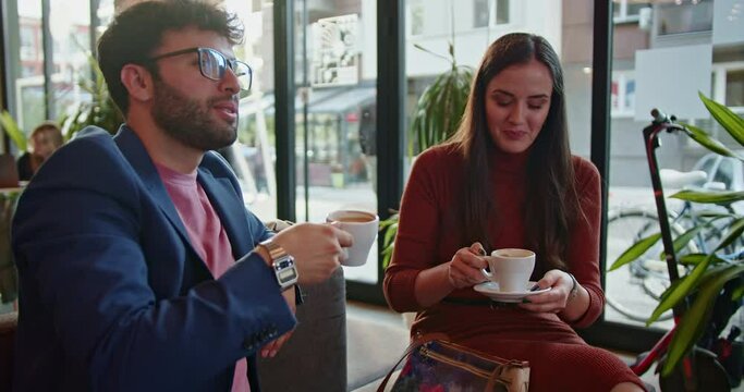 Two professionals engaging in a friendly conversation over coffee in a stylish, modern cafe with natural light and urban views.