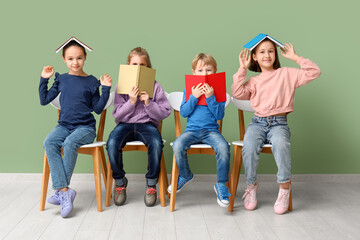 Little children reading books while sitting on chairs near green wall