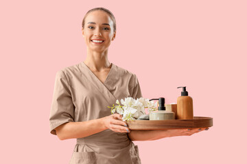 Portrait of female massage therapist holding tray with spa supplies and flowers on pink background