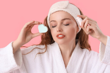 Beautiful woman in bath headband with heart-shaped sponge and makeup foundation on pink background,...
