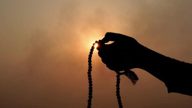 Silhouette Hand of Muslim man carrying tasbih and praying with sunset Background. 