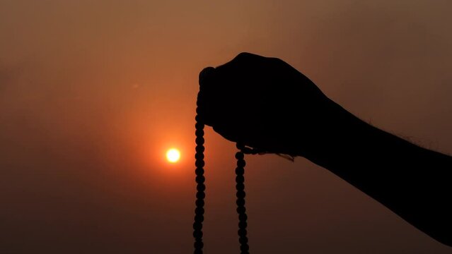 Silhouette Hand of Muslim man carrying tasbih and praying with sunset Background. 