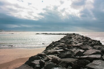 A scenic view of a rocky beach with a stone breakwater in the foreground made of large, rough stones, contrasting with the small stone beach and gray cloudy sky in the background.