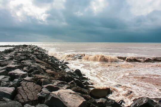 A wide shot of a rocky shore with a stormy sea under a dramatic sky and cloudscape. The water is dark blue and the waves are whitewash, crashing against the stone embankment.