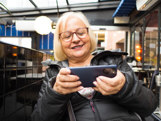gamer grandmother smiling playing on the mobile phone on a restaurant terrace - 780151120